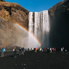Turyści pod wodospadem Skógafoss na Islandii, podziwiający tęczę. Wspaniałe widoki, z jakimi biurem na Islandię warto się wybrać.