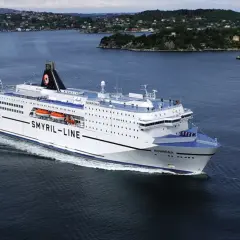 Norröna, prom Smyril Line, płynie przez spokojne wody. W tle widać zielone wzgórza i zabudowania. Idealny widok na podróż na Wyspy Owcze.