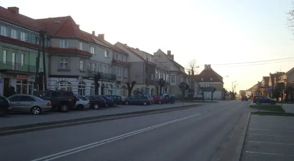 Ulica w miasteczku, gdzie są Wydminy. Rząd kamienic z czerwonymi dachami, zaparkowane samochody i droga.