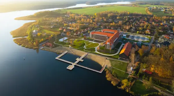 Widok z lotu ptaka na kompleks Lake Hill Mazury z jeziorem, plażą, molo i budynkami w jesiennej scenerii.