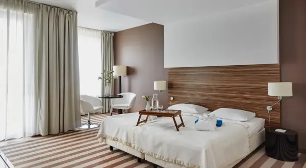 Przytulny pokój hotelowy z dwoma łóżkami, stolikiem i fotelami, ozdobiony drewnianym zagłówkiem i pasiastym dywanem.