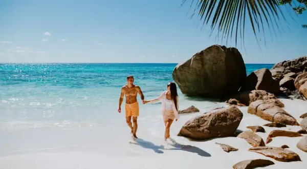 Para idealny czas na podróż na Seszele, para spaceruje po rajskiej plaży z turkusową wodą i skałami.