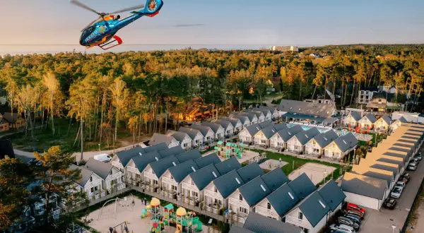 Widok z lotu ptaka na holiday park z domkami, placem zabaw i basenem, a nad nim latający helikopter.