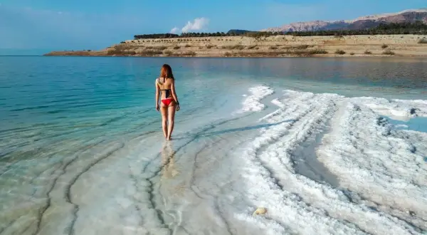Kobieta w czerwonym bikini spaceruje po krystalicznej plaży Morza Martwego w Jordanii.