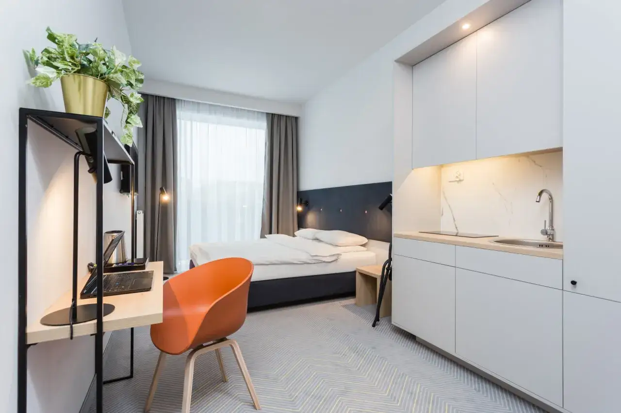 Nowoczesny pokój w hotelu Platinum Residence: łóżko, biurko z laptopem, aneks kuchenny i pomarańczowe krzesło.
