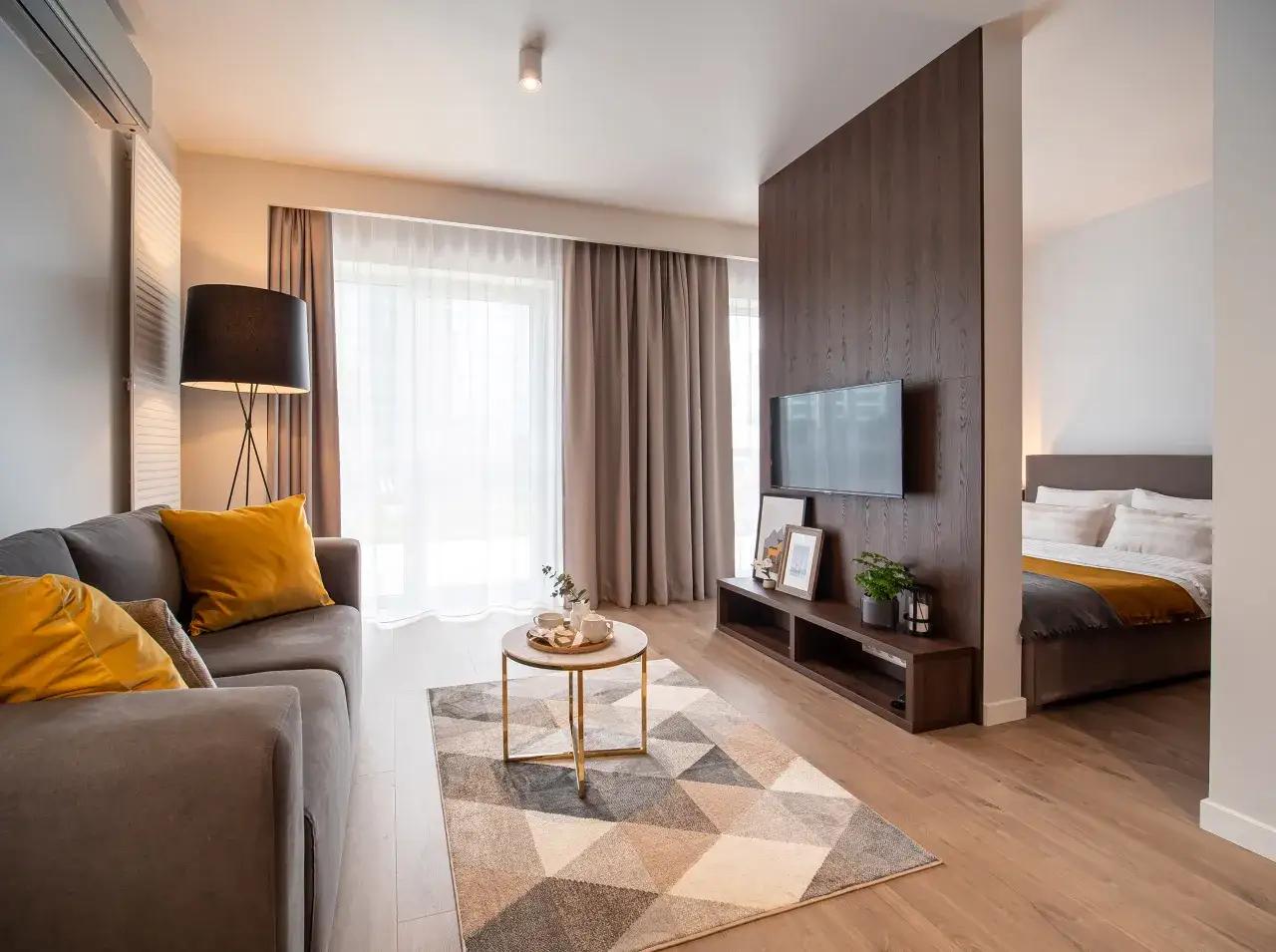 Nowoczesny apartament w hotelu Platinum Residence: salon z szarą kanapą i żółtymi poduszkami, stolik kawowy, dywan, TV, sypialnia z łóżkiem.