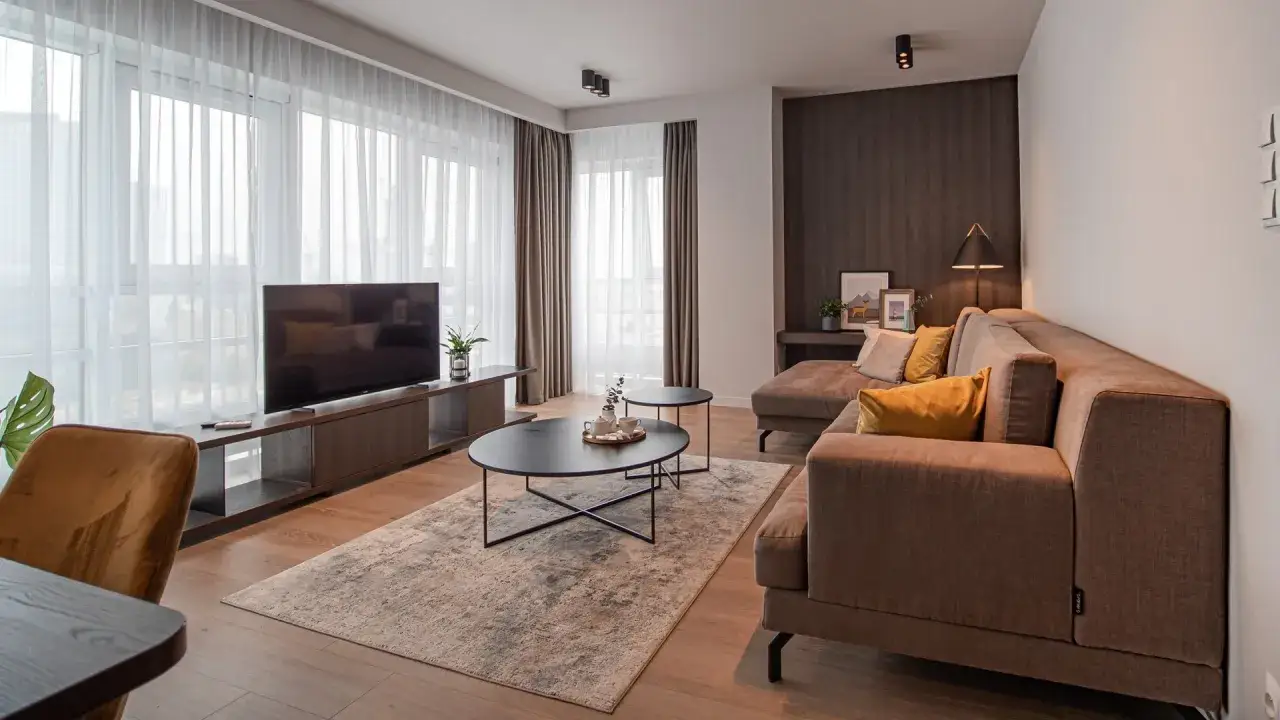 Nowoczesny salon w hotelu Platinum Residence. Brązowa sofa, stoliki kawowe, telewizor i duże okna z zasłonami.